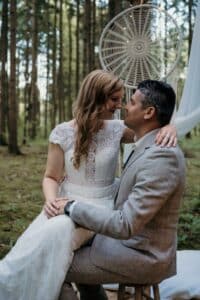 Ein Paar in Hochzeitskleidung sitzt eng beieinander in einer Waldlandschaft. Die Frau in einem weißen Spitzenkleid sitzt auf dem Schoß des Mannes; er trägt einen hellgrauen Anzug. Ein großer, dekorativer Traumfänger, der die Magie ihrer Hochzeit einfängt, hängt hinter ihnen an einem Ast.