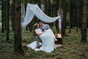 Ein Paar im Hochzeitskleid küsst sich unter einem weißen, drapierten Baldachin in einem Wald. Traumfänger und Kerzenlaternen verstärken die romantische, rustikale Atmosphäre. Die Braut sitzt auf dem Schoß des Bräutigams, umgeben von hohen Bäumen und üppigem Grün – die Essenz einer intimen Hochzeitskulisse.