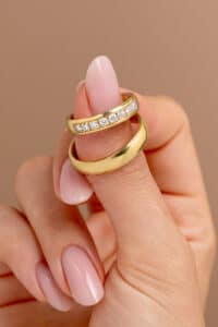 Eine Hand mit sorgfältig manikürten Nägeln hält zwei goldene Ringe, perfekte Symbole der Liebe und Verbundenheit. Ein Ring ist schlichtes Gold, der andere mit kleinen Diamanten verziert. Vor einem sanften braunen Hintergrund fangen diese Ringe die Essenz einer Hochzeit ein und werten jede Hochzeitsplanung auf.