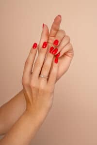 Nahaufnahme der Hände einer Person mit rot lackierten Nägeln, perfekt für eine Hochzeit. Eine Hand trägt einen einfachen Diamantring am Ringfinger, der das Heiraten symbolisiert. Der Hintergrund ist in einem warmen Beige gehalten.