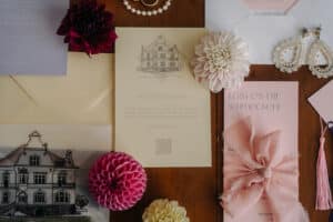 Eine Sammlung von Hochzeitspapeterie auf einem Holztisch, unverzichtbar für jede anspruchsvolle Hochzeitsplanung. Sie umfasst Einladungen mit Abbildungen eines Gebäudes, dekorativen Blumen und einer rosa Schleife. Edle Ohrringe und Umschläge runden das romantische Hochzeitsthema ab.