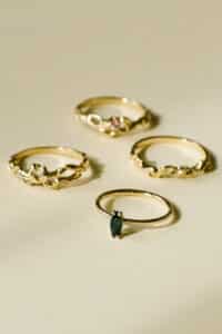 Vier exquisite Goldringe sind auf einer hellen Oberfläche angeordnet, perfekt für Ihre Hochzeitsplanung. Jeder Ring weist ein einzigartiges Design auf, wobei einer einen dunkelgrünen Edelstein aufweist und andere mit zarten Details und kleinen dekorativen Elementen geschmückt sind.