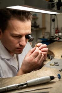 Ein Juwelier, vertieft in seine Hochzeitsplanung, untersucht einen Ring durch eine Lupe. Er trägt einen weißen Kittel und arbeitet unter einer hellen Schreibtischlampe an einer überfüllten Werkbank, umgeben von Werkzeugen und anderen Ringen.