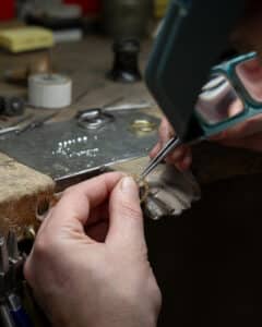 Inmitten einer überfüllten Werkstatt verwendet ein sorgfältiger Handwerker ein Vergrößerungswerkzeug, um ein feines Schmuckstück zu perfektionieren, das vielleicht Teil des besonderen Tages einer Person sein soll. Verstreute Werkzeuge und Materialien verleihen eine Aura der Kreativität, als ob sie sich auf einen so kostbaren Moment wie eine Hochzeit vorbereiten würden.