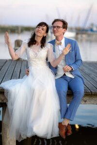 Braut und Bräutigam sitzen während ihrer intimen Hochzeit auf einem Holzsteg am Wasser. Die Braut trägt ein weißes Kleid mit Spitzendetails und der Bräutigam einen blauen Anzug mit Fliege. Beide lächeln, wobei die Braut spielerisch ihre Hände hebt und damit die Freude des Heiratens verkörpert.