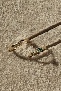 Zwei goldene Ringe werfen Schatten auf eine strukturierte Sandoberfläche, perfekte Symbole für eine Hochzeit. Ein Ring ist schlicht, während der andere mit einem kleinen grünen Edelstein versehen ist, der die Eleganz einer Hochzeit verkörpert.