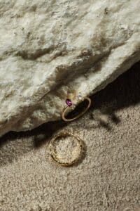 Zwei goldene Ringe auf einer strukturierten Oberfläche: einer mit einem glatten Band, das klare und rote Edelsteine enthält, der andere mit einem strukturierten Band. Sie liegen auf einem rauen Steinhintergrund und werfen weiche Schatten – perfekte Symbole für diejenigen, die ihre Hochzeit planen.