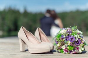 Nahaufnahme von beigen High Heels und einem bunten Blumenstrauß auf einer Holzoberfläche. Im unscharfen Hintergrund sitzt ein Paar zusammen, mit viel Grün und blauem Himmel im Hintergrund, und genießt seine Hochzeitsplanung, während sie sich auf ihre Hochzeit vorbereiten.
