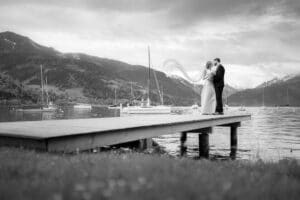 Ein Paar umarmt sich auf einem Holzsteg an einem ruhigen See, umgeben von Bergen und Segelbooten. Der Schleier der Braut weht im Wind und verkörpert die Romantik ihrer Hochzeit. Die Szene ist in Schwarzweiß festgehalten und schafft eine ruhige Atmosphäre, die perfekt ist, um sich an diesen besonderen Tag zu erinnern.