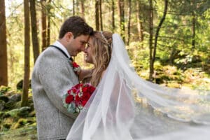 In einem ruhigen Wald steht ein Paar im Hochzeitskleid Stirn an Stirn. Die Braut, die einen Strauß roter Rosen hält und einen wallenden Schleier trägt, verkörpert die Essenz der Hochzeit. Sonnenlicht fällt durch die Bäume und verstärkt die romantische Atmosphäre, während sie ihre Entscheidung zu heiraten feiern.