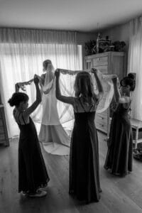 Eine Braut steht in einem fließenden Kleid und Schleier neben einem Fenster und verkörpert die Schönheit einer Hochzeit. Vier Brautjungfern in passenden Kleidern halten den Schleier hoch und schaffen eine dramatische, elegante Szene in einem sanft beleuchteten Raum mit Holzmöbeln – ein unvergesslicher Moment ihrer sorgfältigen Hochzeitsplanung.