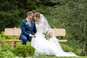 Braut und Bräutigam sitzen eng beieinander auf einer Holzbank in einer üppigen grünen Umgebung und verkörpern den perfekten Moment ihrer Hochzeit. Sie trägt ein weißes Kleid und einen Schleier mit einem Blumenstrauß, während er elegant in seinem blauen Anzug aussieht. Sie berühren liebevoll ihre Stirn, umgeben von Grün.