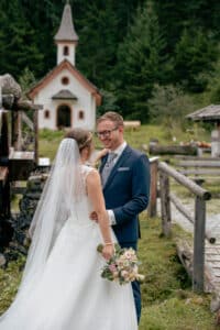 Braut und Bräutigam stehen zusammen im Freien in der Nähe einer kleinen Kapelle und feiern ihre Hochzeit. Die Braut trägt ein weißes Brautkleid und einen Schleier und hält einen Blumenstrauß, während der Bräutigam einen blauen Anzug und eine Brille trägt. Sie lächeln sich an, umgeben von üppigem Grün und der Freude des Heiratens.