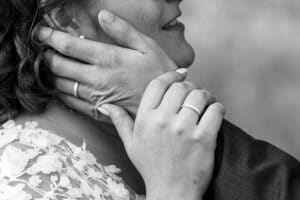 Eine Nahaufnahme in Schwarzweiß von zwei Menschen, die sich während ihrer Hochzeit umarmen. Eine Person hält sanft das Gesicht der anderen und zeigt dabei ihre Eheringe. Die Hand der anderen Person berührt den Hals der Person. Der Hintergrund ist leicht verschwommen, was die Intimität dieses wunderschönen Moments der Hochzeit hervorhebt.