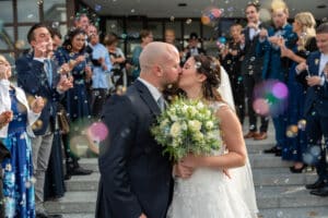 Braut und Bräutigam küssen sich während ihrer Hochzeit auf einer Treppe, während die Gäste um sie herum Seifenblasen pusten. Die Braut hält einen Strauß weißer und grüner Blumen, während Gäste in Abendgarderobe auf beiden Seiten stehen und zur festlichen Atmosphäre ihres wunderschön geplanten Tages beitragen.