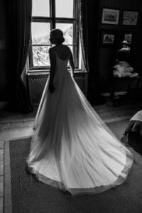 Eine Braut in einem langen, fließenden Kleid steht am Fenster und denkt über ihre Hochzeit nach. Die Schleppe des Kleides bedeckt elegant den Holzboden. In dem schwach beleuchteten Raum hängen gerahmte Bilder an den Wänden und auf einem Regal liegt ein Stapel Handtücher, was inmitten der Hektik der Hochzeitsplanung Ruhe ausstrahlt.