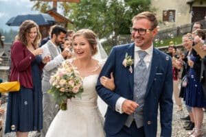 Eine fröhliche Braut und ein fröhlicher Bräutigam gehen Arm in Arm, umgeben von lächelnden Gästen, die Seifenblasen pusten, bei ihrer Hochzeit. Die Braut hält einen Strauß rosa und weißer Blumen, während der Bräutigam einen blauen Anzug trägt. Die Kulisse scheint eine Feier im Freien voller Liebe und Lachen zu sein.