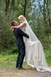 Vor der Kulisse eines üppigen, grünen Waldes hebt ein Bräutigam im dunklen Anzug seine Braut in ihrem weißen Hochzeitskleid mit langem Schleier hoch. Sie lächeln einander an und verkörpern die Freude und Romantik ihres Hochzeitstages.