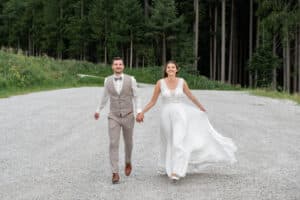 Eine lächelnde Braut und ein lächelnder Bräutigam halten sich an den Händen, während sie auf einem Kiesweg gehen und so die Freude ihrer Hochzeit zum Ausdruck bringen. Die Braut trägt ein fließendes weißes Kleid und der Bräutigam ist mit einer beigefarbenen Weste und Hose sowie einer Fliege bekleidet. Sie sind von Grün und hohen Bäumen umgeben, was ihrem perfekten Tag einen Hauch von Natur verleiht.