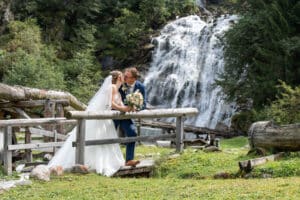 Braut und Bräutigam küssen sich vor einem Wasserfall, umgeben von üppigem Grün. Die Braut trägt ein weißes Kleid und einen Schleier und hält einen Blumenstrauß, während der Bräutigam einen Anzug trägt. Sie stehen auf einer kleinen Holzbrücke und schaffen so die perfekte romantische Kulisse für ihre Hochzeit im Freien.