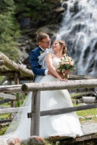 Braut und Bräutigam umarmen sich auf einer Holzbrücke mit einem Wasserfall im Hintergrund und fangen so die Essenz ihrer Hochzeit ein. Die Braut hält einen Blumenstrauß, während sie sich anlächeln, inmitten einer üppigen, grünen Außenumgebung, die perfekt zum Heiraten ist.