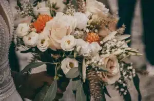 Eine Nahaufnahme einer Hand, die einen Brautstrauß hält, perfekt für alle, die sich auf Hochzeitsplanung konzentrieren. Er besteht aus weißen und pfirsichfarbenen Rosen, orangefarbenen Blüten und üppigem Grün. Das komplizierte Design und die Vielfalt der Texturen ergeben ein elegantes Arrangement für den Hochzeitstag.