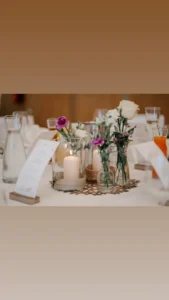 Ein wunderschön arrangierter Tischschmuck mit Kerzen, gemischten Blumen in Glasvasen und Wasserkrügen schafft die Atmosphäre für eine warme und einladende Hochzeit. Elegant gedeckt mit Menüs und Gläsern ist es perfekt für diejenigen, die bei ihrer Hochzeitsplanung besondere Momente schätzen.