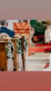 Eine Szene aus einer Hochzeitszeremonie zeigt den Innenraum einer Kirche mit Holzbänken, die mit weißen und grünen Blumendekorationen geschmückt sind. Die Menschen sitzen in formeller Kleidung und verkörpern damit die Essenz einer sorgfältigen Hochzeitsplanung, während vorne in der Nähe des Altars eine verschwommene Gestalt steht.