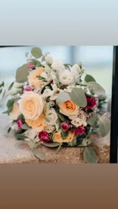 Ein lebendiger Strauß mit pfirsichfarbenen Rosen, weißen Blumen und kleinen rosa Blüten, akzentuiert durch üppiges Grün, steht auf einer Steinoberfläche. Dieses elegante Arrangement ist perfekt für die Hochzeitsplanung und schafft ein frisches Ambiente, das sich ideal für Hochzeitsfeiern eignet.