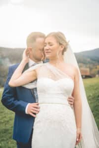 Ein frisch vermähltes Paar steht im Freien und feiert seine Hochzeit. Die Braut trägt ein weißes, einschultriges Kleid und einen Schleier, während der Bräutigam einen blauen Anzug trägt. Sie posieren eng beieinander, mit einem offenen Feld und Hügeln im Hintergrund – und fangen einen perfekten Moment ihres besonderen Tages ein.
