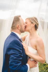 Ein Paar küsst sich an seinem Hochzeitstag. Die Braut trägt ein fließendes weißes Kleid und einen Schleier und der Bräutigam einen blauen Anzug. Eingehüllt in weichen, weißen Stoff fängt dieser intime Moment die Essenz ihrer Hochzeit ein – wahrlich eine verträumte Atmosphäre, die einem so geschätzten Fest würdig ist.