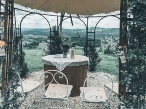 Eine rustikale Außenkulisse, perfekt für die Hochzeitsplanung, mit Vintage-Metallstühlen und einem Tisch unter einem kunstvollen Pavillon. Auf dem mit Spitze bedeckten Tisch steht eine Kerze, während die malerische Aussicht auf sanfte Hügel und Felder unter einem bewölkten Himmel eine idyllische Kulisse für jedes Paar schafft, das sich auf seine Hochzeit vorbereitet.