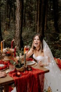 Eine Braut in einem weißen Kleid und einem transparenten Schleier sitzt an einem rustikalen Tisch im Wald und verkörpert die Essenz der Hochzeitsplanung. Der Tisch ist mit rotem Stoff, Blumen, Kerzen und gewebten Dekorationen geschmückt. Sie hält ein Weinglas und lächelt freudig inmitten des üppigen Grüns.