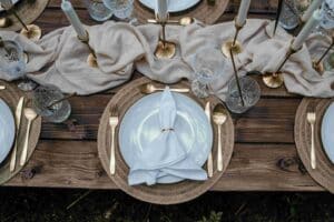 Ein elegant gedeckter rustikaler Tisch mit Holzoberfläche verfügt über einen weißen Teller, goldenes Besteck und eine gefaltete weiße Serviette. Kristallgläser und hohe goldene Kerzenständer mit weißen Kerzen sorgen für Eleganz, begleitet von einem Tischläufer aus elfenbeinfarbenem Stoff – perfekt für jede Hochzeitsfeier.