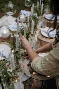 Eine Person arrangiert weiße Kerzen auf einem wunderschön dekorierten Esstisch im Freien, der mit Grünpflanzen und eleganten Gedecken geschmückt ist. Die Atmosphäre ist rustikal und charmant, perfekt für eine romantische Hochzeit oder eine besondere Feier.
