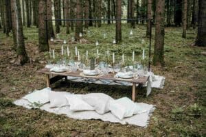 Eine rustikale Hochzeitstafel im Freien in einem Wald präsentiert einen Holztisch mit weißen Kerzen, Grünpflanzen und Geschirr. Hängende Lichter funkeln darüber. Weiße Kissen sind auf einem Teppich um den Tisch herum inmitten der Bäume platziert und schaffen eine zauberhafte Atmosphäre für eine Hochzeit unter den Sternen.