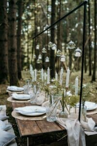 Eine rustikale Tischdekoration für eine Hochzeit im Freien im Wald besteht aus einem langen Holztisch, der mit weißen Kerzen, Farnen in Glasvasen und kleinen Glasornamenten geschmückt ist, die über dem Tisch hängen. Tischsets und Kissen in neutralen Farben dienen als Sitzgelegenheit und Dekoration.