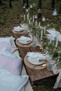 Eine rustikale Tischdekoration im Freien in einem Wald strahlt Hochzeitscharme aus. Sie besteht aus einem Holztisch mit weißen Tellern, goldenem Besteck, Kristallgläsern und grünen Blattwerk-Mittelstücken. Hängende Kerzenhalter aus Glas verleihen eine magische Note, perfekt für jede Hochzeit. Weiche blaue Kissen dienen als Sitzgelegenheiten.