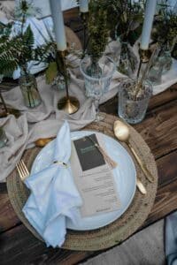 Ein rustikales Tischgedeck für eine Hochzeit besteht aus einem weißen Teller, goldenem Besteck und einer blauen Serviette. Auf dem Teller liegt anmutig ein Menü. Der Tisch ist elegant mit grünem Laub, Kerzenleuchtern und Glaswaren auf einer Holzoberfläche mit einem Tischläufer aus beigem Stoff geschmückt.