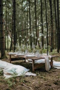 Eine rustikale Tischdekoration im Freien in einem Wald, perfekt für die Hochzeitsplanung, besteht aus einem Holztisch, der mit grünem Laub, weißen Kerzen und hängenden Glaskugeln geschmückt ist. Weiße Sitzkissen sind rundherum verteilt und schaffen eine elegante und natürliche Atmosphäre, ideal für die Feier Ihrer Hochzeit.
