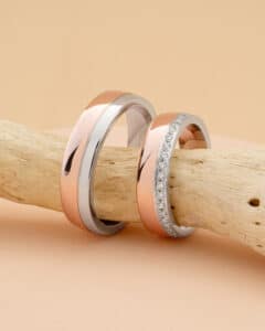 Zwei elegante Ringe ruhen auf einem Stück Treibholz vor einem beigen Hintergrund, perfekte Symbole für jeden, der gerade mitten in der Hochzeitsplanung steckt. Ein Ring hat ein poliertes Rosé- und Silberband, während der andere Rosé- und Silbertöne mit einer Reihe kleiner, funkelnder Steine hat.
