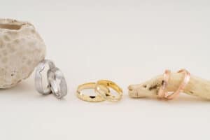 Eine Präsentation verschiedener Ringe auf neutralem Hintergrund verströmt die Essenz des Heiratens, mit Paaren von Silber-, Gold- und Roségoldringen neben rustikalen Strandelementen wie Korallen und Treibholz, wodurch eine natürliche, elegante Präsentation entsteht, die perfekt für die Hochzeitsplanung ist.