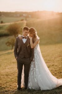 Ein Paar steht bei Sonnenuntergang eng beieinander auf einem sonnenbeschienenen Feld und fängt die Essenz ihrer Hochzeit ein. Die Frau in einem weißen Spitzenbrautkleid lehnt sich sanft an die Schulter des Mannes. Er trägt einen braunen Anzug mit Weste und Fliege. Im Hintergrund sind sanfte Hügel und Bäume zu sehen.
