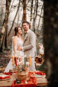 Ein Paar in Hochzeitskleidung steht lächelnd in einer Waldlandschaft und verkörpert die Essenz einer Hochzeit. Die Braut trägt ein weißes Kleid und der Bräutigam einen hellen Anzug. Im Vordergrund ist ein Tisch elegant mit Kerzen und herbstlichen Arrangements dekoriert – perfekt für jede Hochzeitsplanung.