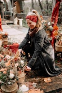 Eine Frau in einem schwarzen Kleid und einem roten Stirnband arrangiert Blumendekorationen auf einem Holztisch im Freien und bereitet so die Bühne für eine bezaubernde Hochzeit. Umgeben von herbstlich gefärbten Blättern, weißen Rosen und geflochtenen Körben strahlt die Atmosphäre warme, rustikale Eleganz aus, perfekt zum Heiraten.
