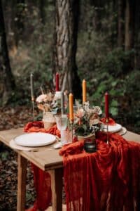 Eine rustikale Außenkulisse, die sich perfekt für ein gemütliches Herbstessen oder eine bezaubernde Hochzeit eignet, besteht aus zwei weißen Tellern, bunten Kerzen und Trockenblumen. Ein roter Stoff hängt über dem Holztisch und harmoniert mit der Waldkulisse. Ideal für alle, die eine einzigartige Hochzeitsplanung planen.