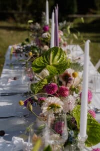 Ein wunderschön gedeckter Tisch im Freien mit einem eleganten weißen Tuch ist mit großen weißen und lavendelfarbenen Kerzen und einem üppigen Blumenschmuck mit grünen Blättern, rosa und weißen Blumen geschmückt. Die Hochzeitskulisse strahlt eine frische und lebendige Atmosphäre aus, perfekt für eine Hochzeit unter freiem Himmel.