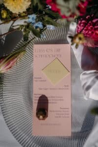 Eine Menükarte mit Blumenschmuck liegt auf einem Glasteller und wurde für eine Hochzeitsfeier entworfen. Auf der Karte steht eine deutsche Liste der Gänge des Menüs, darunter Suppe, Hauptgericht und Nachtisch. Leuchtend rosa und weiße Blumen zieren das elegante Arrangement.