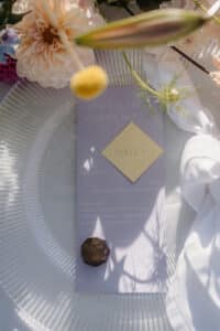 Ein zartes Tischgedeck für eine Hochzeit besteht aus einem Glasteller, einem Blumenarrangement und einer lavendelfarbenen Menükarte mit dem Namen „Patrick“ auf einem beigen Quadrat. Auf dem Teller liegt eine Pralinenkugel, die dieser wunderschön geplanten Szene eine elegante Note verleiht.