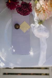 Auf einem durchsichtigen Glasteller liegt eine lavendelfarbene Menükarte, die mit einem quadratischen Etikett in Schokoladen- und Goldtönen verziert ist – perfekt für eine Hochzeitsfeier. Elegant neben einer weißen Stoffserviette platziert, ist die Szene mit bunten Blumen geschmückt und verleiht der Hochzeitsplanung Charme.